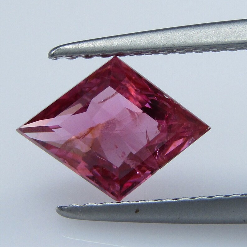 May include: A pink square-shaped gemstone with a faceted cut. The gemstone is held by tweezers.