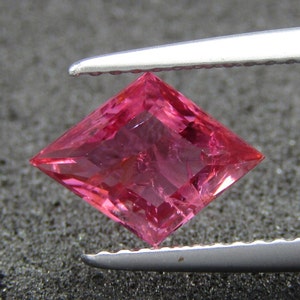 May include: A pink square-shaped gemstone with a faceted cut. The gemstone is held by a pair of tweezers.
