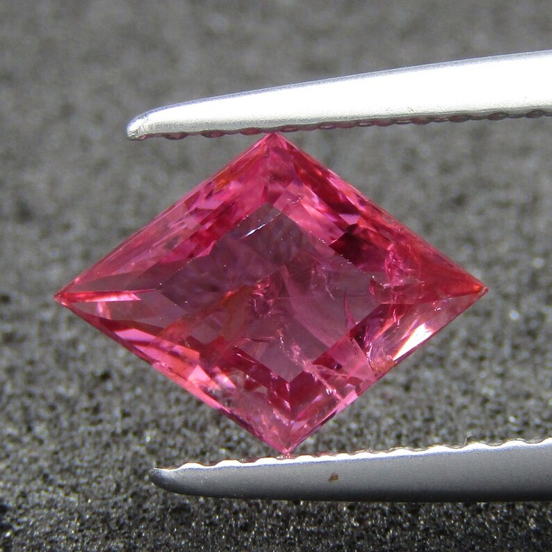 May include: A pink square-shaped gemstone with a faceted cut. The gemstone is held by a pair of tweezers.