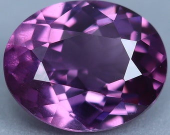 May include: A purple gemstone with a faceted oval cut. The gemstone is set against a gray background.