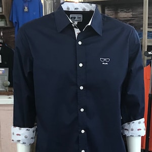 May include: A navy blue long-sleeved button-down shirt with a white collar and cuffs. The shirt has a small white embroidered design of a pair of glasses and a mustache on the front.