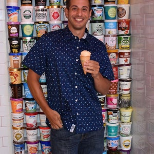 May include: A man wearing a blue patterned shirt and jeans is holding an ice cream cone in front of a wall of ice cream containers. The containers are stacked in rows and feature various flavors and brands.