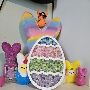 May include: An Easter-themed display featuring a large, white egg-shaped decoration filled with colorful floral confetti. Surrounding the egg are plush bunny figures, a jar with bunny graphics, and a rainbow-colored bunny-shaped pillow.