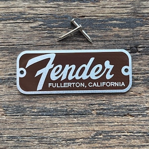 May include: A brown and white Fender guitar headstock logo with the text "Fender Fullerton, California". Two small silver pins are above the logo. The items are on a weathered wooden surface.