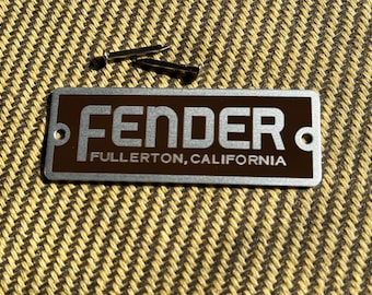 May include: A rectangular brown and silver Fender badge with the text "FENDER" and "FULLERTON, CALIFORNIA" in white. Two small silver rivets are above the badge. The background is a textured tan and yellow fabric.
