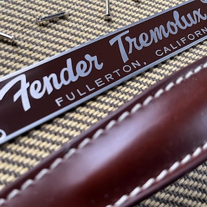 May include: A silver metal plate with the text "Fender Tremololux Fullerton, California" on a brown background. The plate is attached to a brown leather strap with white stitching.