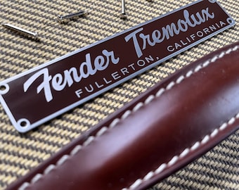 May include: A silver metal plate with the text "Fender Tremololux Fullerton, California" on a brown background. The plate is attached to a brown leather strap with white stitching.