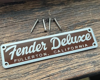 May include: A silver metal plate with a brown background. The plate has the text "Fender Deluxe" in white lettering with the words "Fullerton, California" below. The plate has four small holes in the corners.