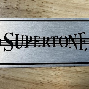 May include: A silver metal plate with a black border and the word "SUPERTONE" in black lettering with musical notes above it.