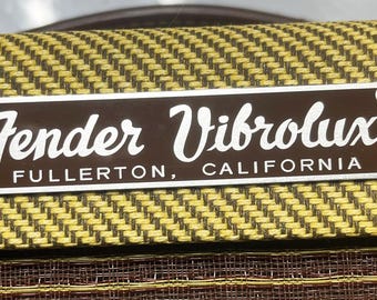 May include: A silver metal plate with a brown background and the text "Fender Vibrolux Fullerton, California" in white lettering.
