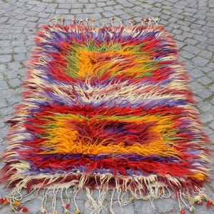 May include: A colorful, hand-woven rug with a shaggy texture. The rug features a geometric pattern in shades of red, orange, yellow, green, purple, and white.