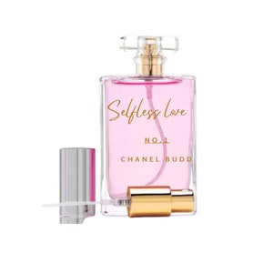 May include: A clear glass bottle of perfume with a gold cap and a gold atomizer. The bottle is filled with a pink liquid and has the words "Selfless Love" and "Chanel Budd" printed on the front. The bottle is sitting on a white background.