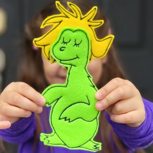 May include: A green felt dinosaur with black outline and closed eyes, and a yellow felt sun shape. The dinosaur is being held up in front of a person.