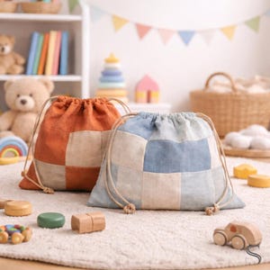 May include: Two drawstring bags, one orange and one blue, with a patchwork design. The bags are made of fabric and have a natural rope drawstring closure. The bags are on a white rug with wooden toys.