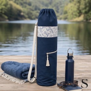 Op de afbeelding: Een marineblauwe yogamat tas met een crèmekleurige band en een bloemenpatroon. Een bijpassende handdoek, waterfles en zonnebril liggen naast de tas. De tas heeft een rits en een kwastje.