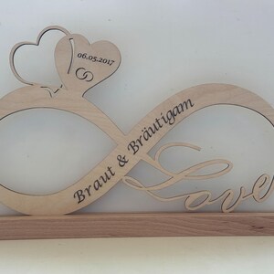 Könnte beinhalten: Holz-Unendlichkeitszeichen mit den Worten "Braut & Bräutigam" und "Love" eingraviert. Zwei Herzen befinden sich oben auf dem Unendlichkeitszeichen mit dem Datum "06.05.2017" eingraviert.