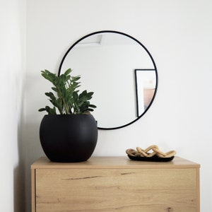May include: A black round mirror with a black frame hangs on a white wall above a light wood dresser. A black planter with a green plant sits on the dresser. A decorative wooden object sits on the dresser.
