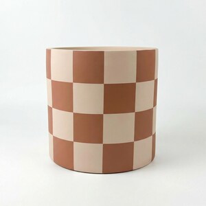 May include: A cylindrical planter with a checkerboard pattern in shades of terracotta and cream. The large, round pot is suitable for indoor or outdoor use. The design is modern and minimalist.