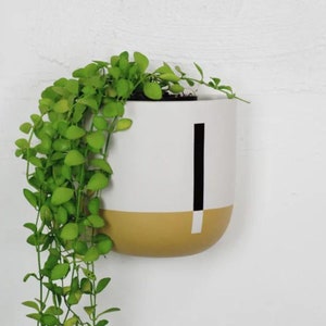 May include: A white and mustard yellow ceramic wall planter with a black stripe design. A trailing green plant is growing out of the planter.