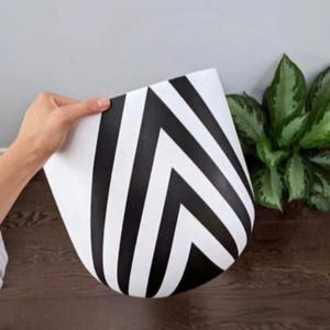 May include: A white planter with a black chevron pattern. The planter is held by a hand, showcasing its design. A green plant is visible in the background, adding a natural element to the scene. The planter's design is modern and geometric.