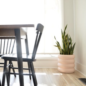 May include: A black dining table with two black chairs and a potted snake plant with a light pink planter. The table and chairs are in a room with a large window and white walls.