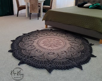 Puede incluir: Una alfombra redonda grande de crochet con un diseño de mandala en tonos grises y rosados. La alfombra está sobre un suelo gris claro en una habitación con una cama y una mesa con sillas.