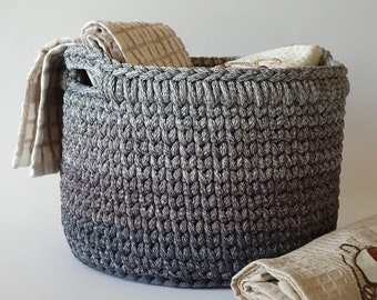 May include: A gray ombre crocheted storage basket with a light gray top and a dark gray bottom. The basket is filled with folded towels and is sitting on a white surface.