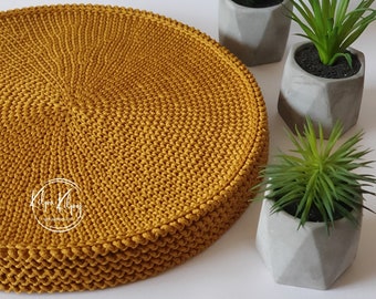 Op de afbeelding: Een ronde, mosterdgele gehaakte onderzetter. De onderzetter is gemaakt van dik garen en heeft een textuur. Hij ligt op een wit oppervlak met drie kleine planten in potten.