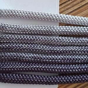 Può includere: Primo piano di diversi fili di corda intrecciata in varie tonalità di grigio e blu scuro. I cordoncini sono etichettati con numeri e testo, tra cui "Nr. 11" e "Nr. 15". I cordoncini sono probabilmente utilizzati per l'artigianato o scopi decorativi.