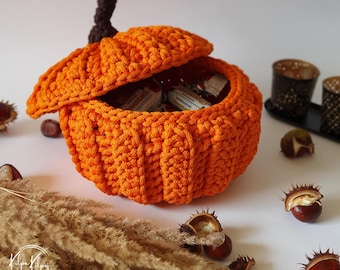 May include: A bright orange crocheted pumpkin-shaped container with a dark brown stem. The lid is slightly ajar, revealing small wrapped items inside. The pumpkin features a ribbed texture created by thick yarn stitches and a rounded base.