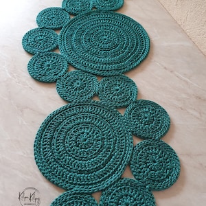 May include: Teal crocheted rug made of multiple circles of varying sizes. The rug is laid out on a light beige surface.