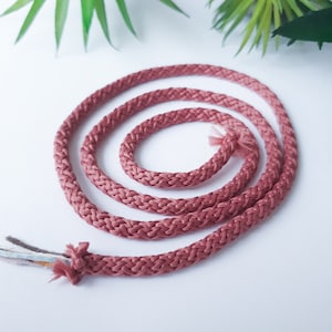 May include: A close-up of a coiled length of pink braided cord. The cord is made of a soft, woven material and has a subtle sheen.