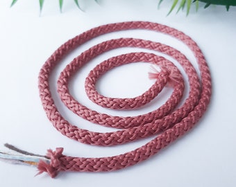 May include: A close-up of a coiled length of pink braided cord. The cord is made of a soft, woven material and has a subtle sheen.