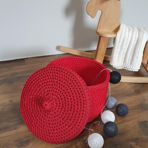 Può includere: Un cesto di vimini rosso lavorato a uncinetto con un coperchio. Il cesto è appoggiato su un pavimento di legno con palline di cotone bianche e grigie sparse intorno.