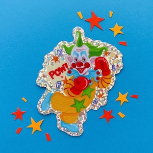 May include: A holographic sticker featuring a clown with green hair, a red nose, and red boxing gloves. The clown wears a yellow and orange striped outfit with a blue collar and shoes. The word POW is printed in red next to the clown.