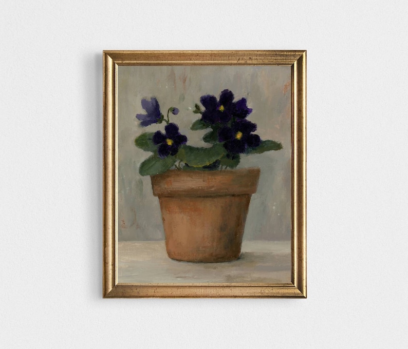 May include: A painting of a potted violet plant with purple flowers and yellow centers. The plant is in a terracotta pot and is sitting on a light gray surface. The painting is framed in a gold frame.