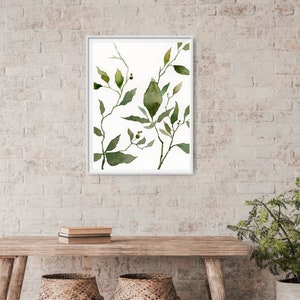 Puede incluir: Una pintura de acuarela enmarcada de hojas verdes y ramas sobre un fondo blanco. La pintura est&aacute; en un marco blanco y est&aacute; colgada en una pared de ladrillo blanco.