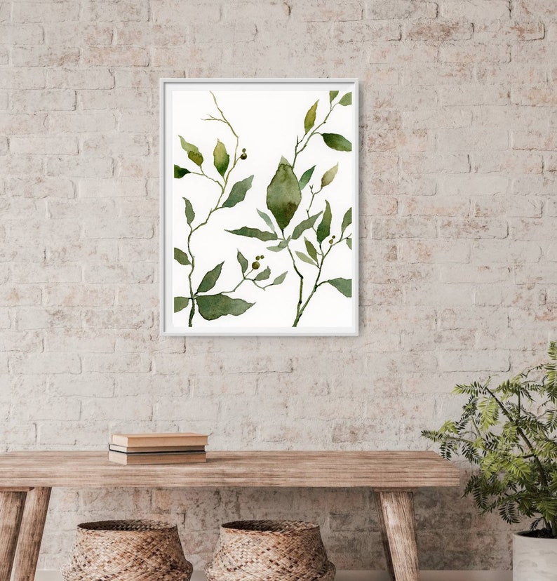 Puede incluir: Una pintura de acuarela enmarcada de hojas verdes y ramas sobre un fondo blanco. La pintura est&aacute; en un marco blanco y est&aacute; colgada en una pared de ladrillo blanco.