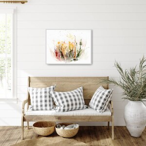K&ouml;nnte beinhalten: Ein Aquarellbild eines Wildblumenfeldes in Gelb-, Gr&uuml;n-, Rot- und Braunt&ouml;nen. Das Bild ist gerahmt und h&auml;ngt an einer wei&szlig;en Wand &uuml;ber einer Holzbank mit Kissen.