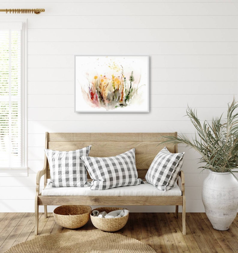 K&ouml;nnte beinhalten: Ein Aquarellbild eines Wildblumenfeldes in Gelb-, Gr&uuml;n-, Rot- und Braunt&ouml;nen. Das Bild ist gerahmt und h&auml;ngt an einer wei&szlig;en Wand &uuml;ber einer Holzbank mit Kissen.