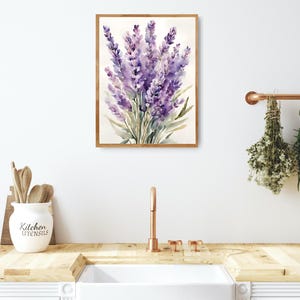 May include: A framed watercolour painting of lavender flowers. The artwork, in shades of purple and green, has a soft, artistic style. The painting is displayed on a white wall, suggesting a home decor theme. The frame is made of wood.