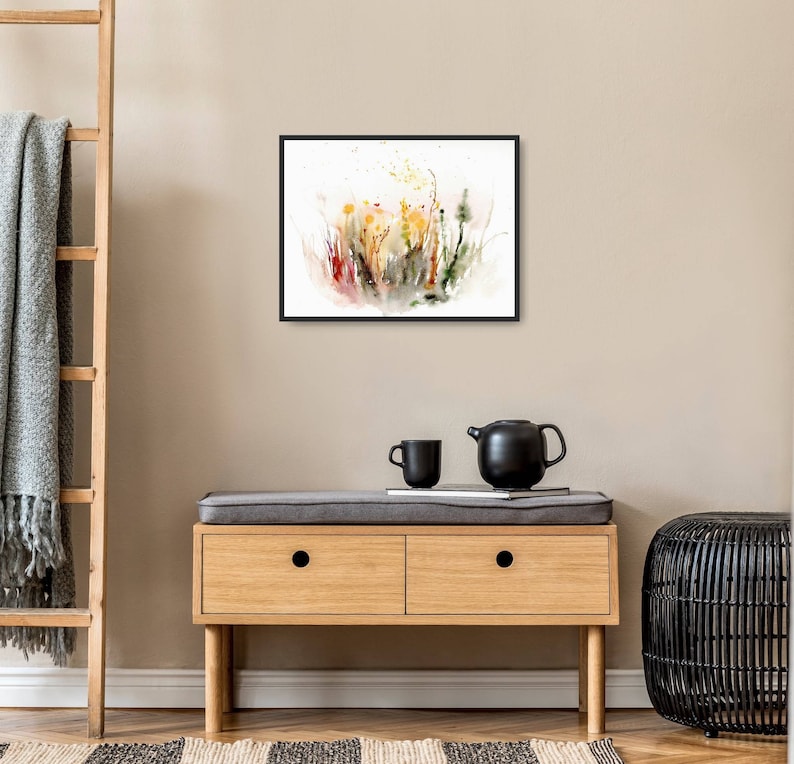 K&ouml;nnte beinhalten: Ein gerahmtes Aquarellbild eines Feldes mit Wildblumen in Gelb-, Orange-, Rot- und Gr&uuml;nt&ouml;nen. Das Bild h&auml;ngt an einer hellbeigen Wand &uuml;ber einer Kommode aus Holz mit zwei Schubladen.
