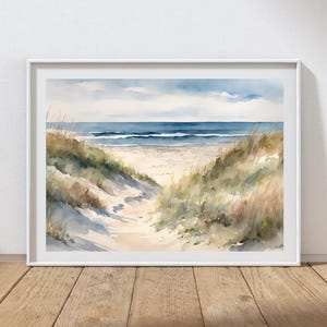 May include: A framed watercolour painting of a beach scene. The artwork shows a sandy path leading to the sea, flanked by dunes and grasses. The sky is a mix of blue and white, and the water is a deep blue.
