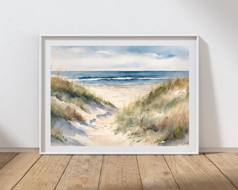 May include: A framed watercolour painting of a beach scene. The artwork shows a sandy path leading to the sea, flanked by dunes and grasses. The sky is a mix of blue and white, and the water is a deep blue.