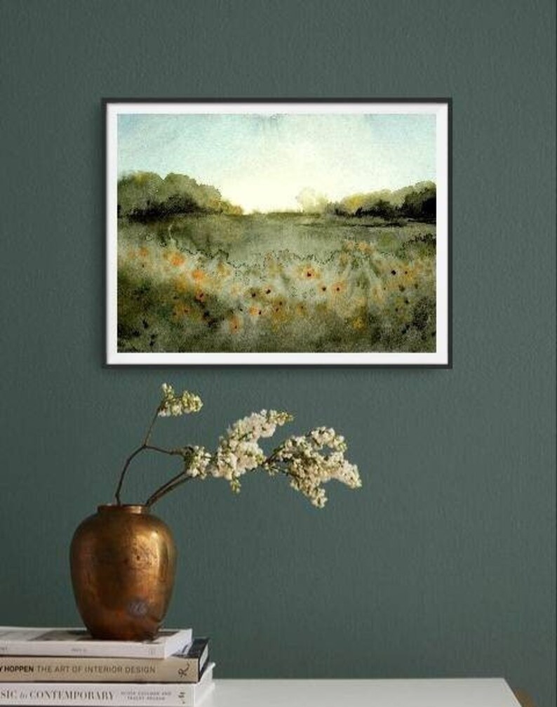 May include: A framed watercolor painting of a field with green grass and a few orange flowers. The painting has a light blue sky with a few white clouds. The painting is framed in a black frame.