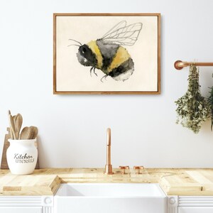 May include: A watercolor painting of a bumblebee with black and yellow stripes and translucent wings. The painting is framed in a light wood frame and is hanging on a white wall.