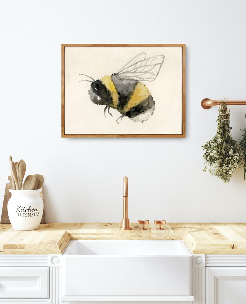 May include: A watercolor painting of a bumblebee with black and yellow stripes and translucent wings. The painting is framed in a light wood frame and is hanging on a white wall.