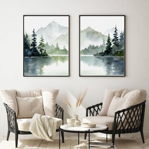 May include: Two framed watercolor paintings of a misty lake scene with mountains and trees in the background. The paintings are in black frames and are hung on a white wall.