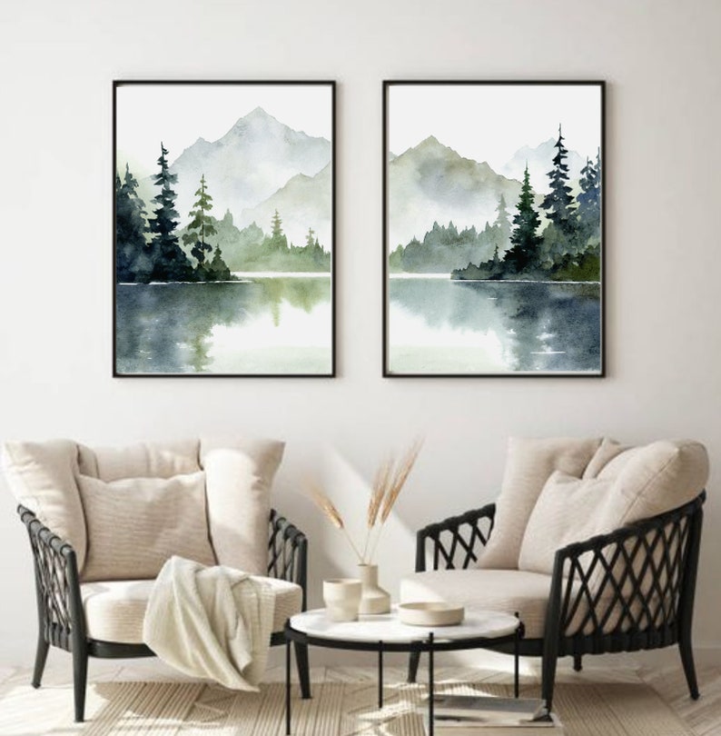 May include: Two framed watercolor paintings of a misty lake scene with mountains and trees in the background. The paintings are in black frames and are hung on a white wall.