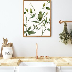 Puede incluir: Una pintura de acuarela enmarcada de hojas verdes y ramas sobre un fondo blanco. La pintura est&aacute; colgada en una pared blanca sobre un fregadero de cocina con un grifo de cobre.